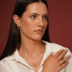 A woman with long dark hair wearing a white collared shirt models gold plated jewelry, including a gold hoop earring, a delicate gold necklace with a green and gold plated pendant, and a gold plated ring with a green stone, against a solid dark reddish-brown background.