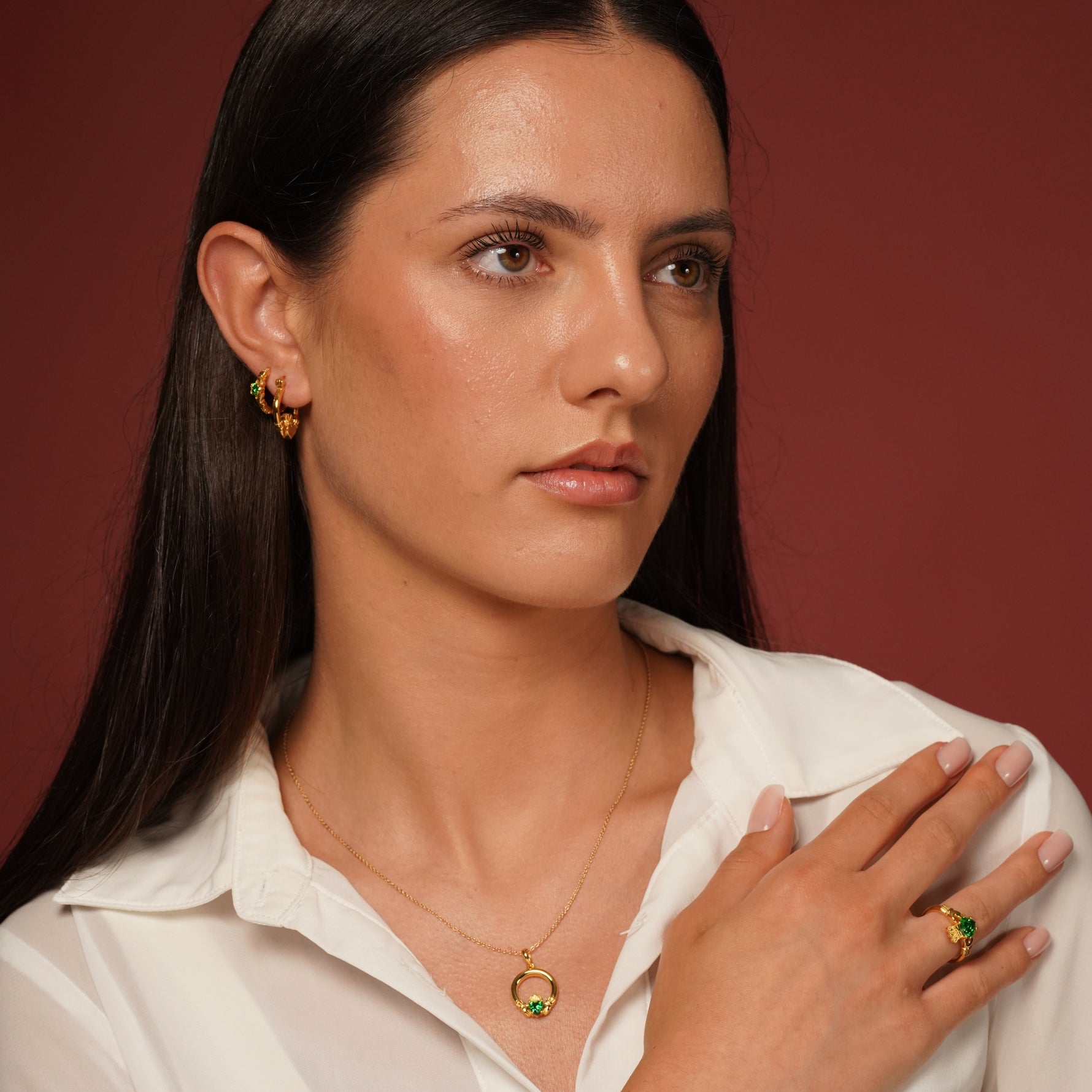 A woman with long dark hair wearing a white collared shirt models gold plated jewelry, including a gold hoop earring, a delicate gold necklace with a green and gold plated pendant, and a gold plated ring with a green stone, against a solid dark reddish-brown background.
