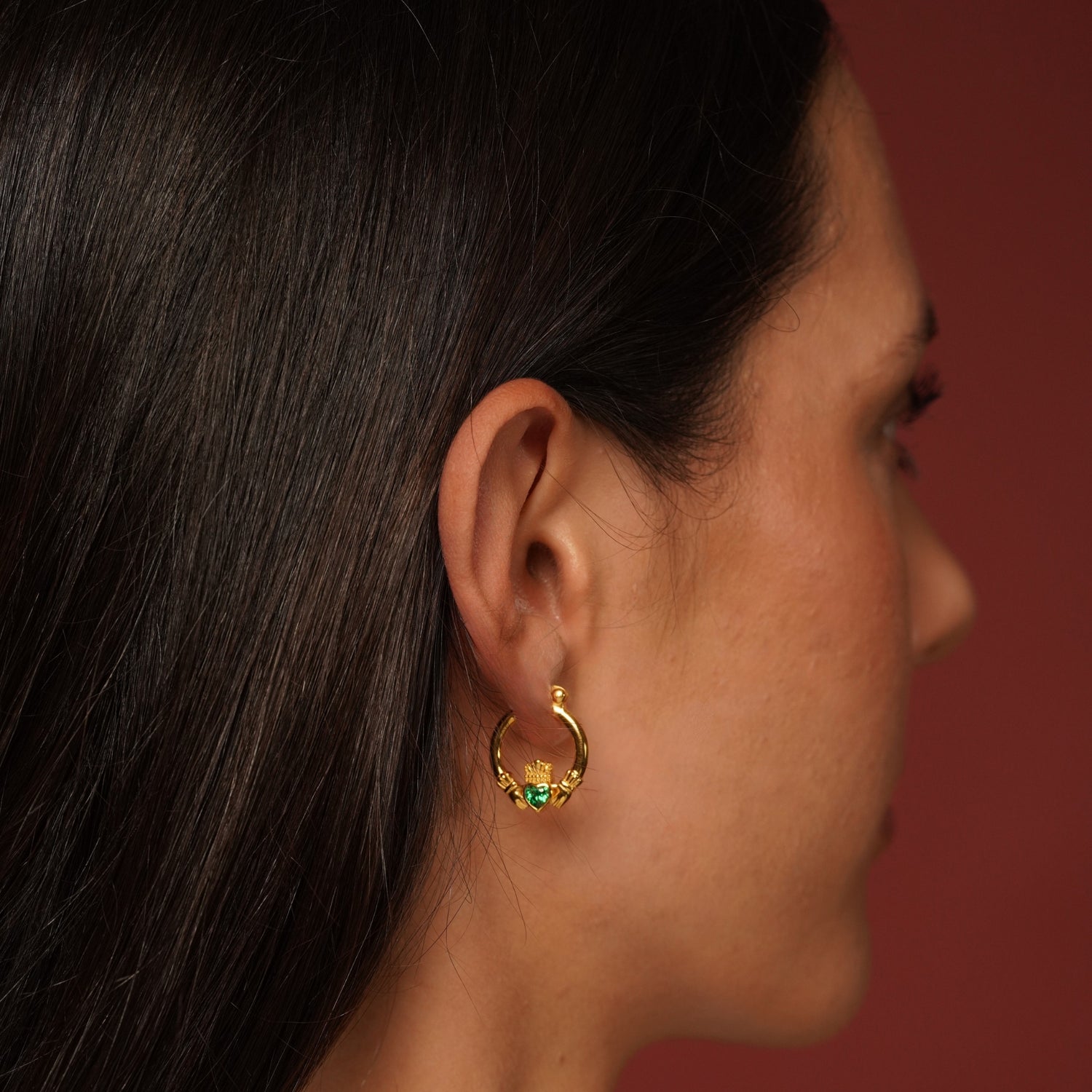 A close-up side profile of a woman with dark hair wearing a small gold plated hoop earring adorned with green and blue stones, set against a solid dark red background.