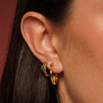 Close-up of a person's ear wearing two gold plated huggie hoop earrings, one adorned with small green gemstones and the other with a textured gold charm.