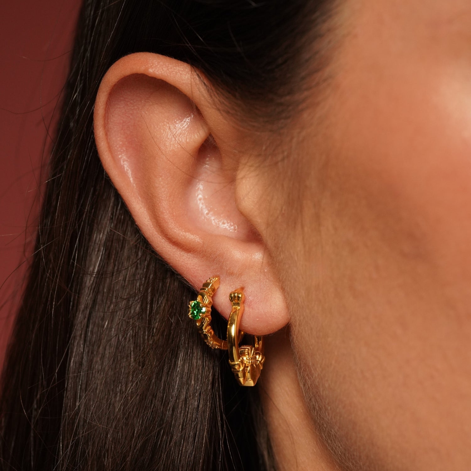 Close-up of a person's ear wearing two gold plated huggie hoop earrings, one adorned with small green gemstones and the other with a textured gold charm.